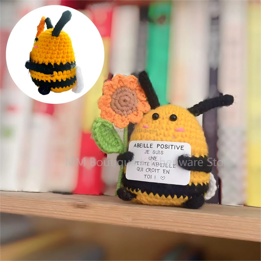 Peluche Abeille Positive au crochet avec tournesol, posée sur une étagère devant des livres, tenant une pancarte motivation “Je suis une petite abeille qui croit en toi”